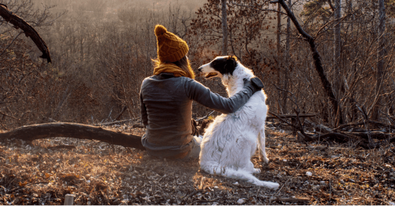 A woman with a brown bobble hat and glove with her arm around a white setter dog with brown ears, sitting on the ground. She has her back turned to the camer and the dog's head is turned looking a her.