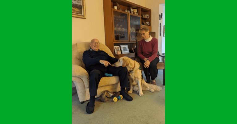 A man sitting in a chair with a golden retriever dog sitting at his feet with his head in his lap. And a woman sitting in a chair next to the dog.