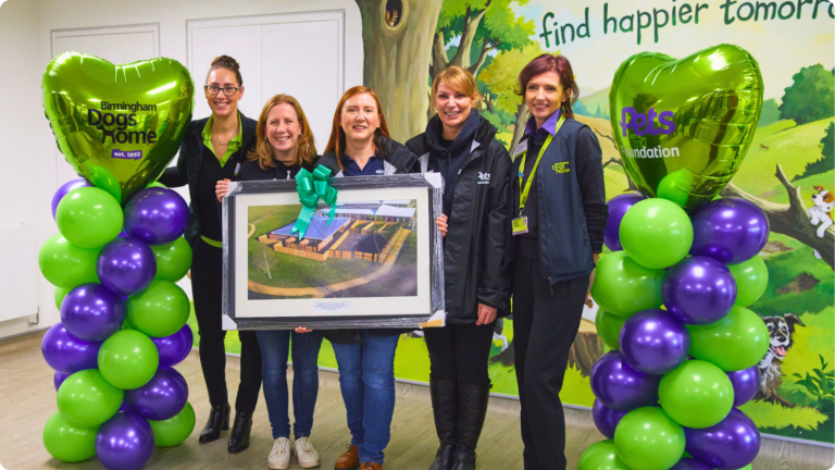 A group of women from Pets Foundation and Birmingham Dogs Home holding a framed picture of the new behavioural unit with green and purple balloons on each side of them.