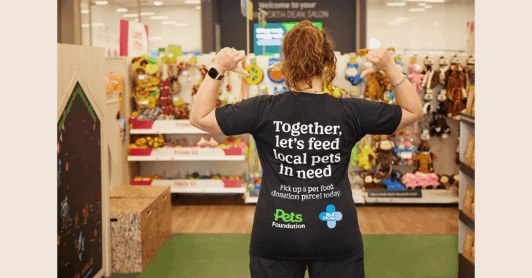A Pets at Home colleague facing away from the camera in store as she's pointing to the back of her shirt that says: Together, let's feed local pets in need. Pick up a pet food donation parcel today. Pets Foundation and Blue Cross logos below the text.