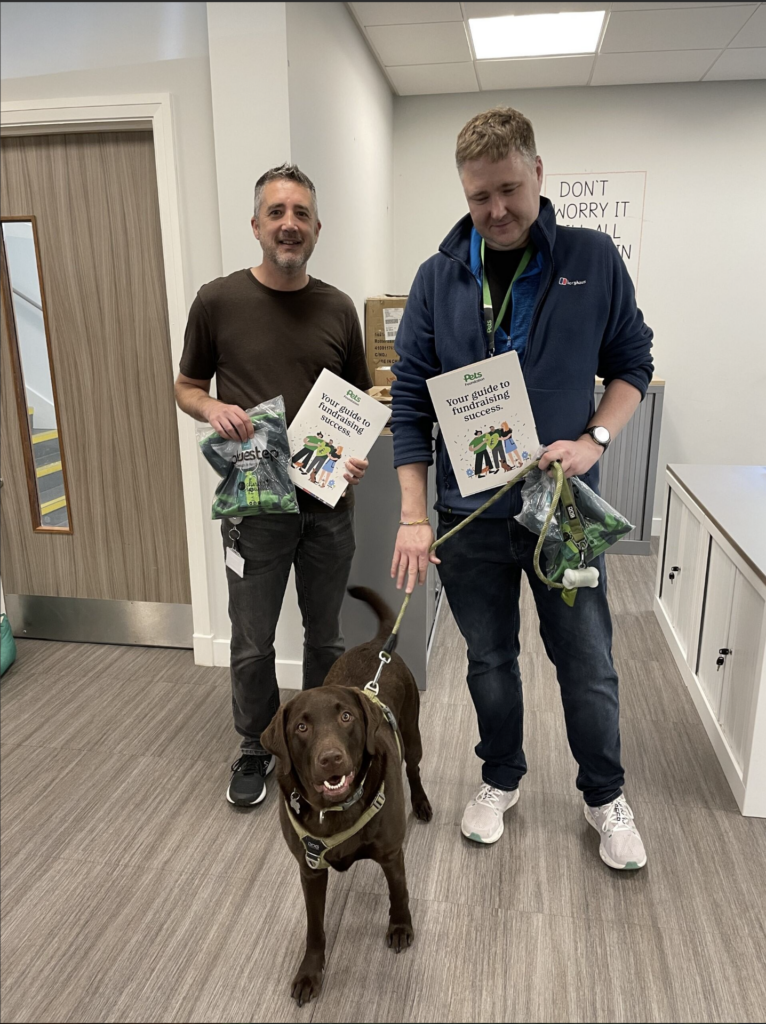 Two men with a brown dog standing in an office holding Pets Foundation fundrasing packs and shirts.
