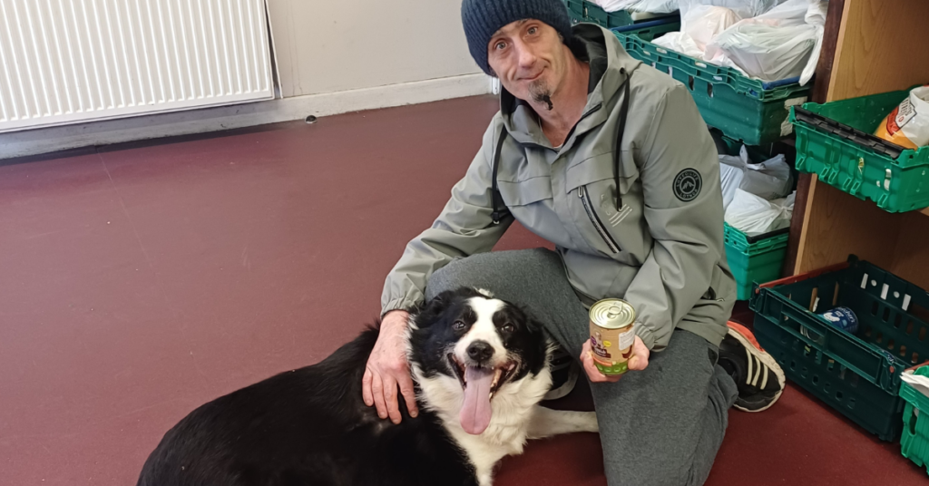 A man sitting in a food bank with a jar of dog food and his black and white dog in his lap.