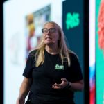 Amy Angus stands on a stage wearing a black Pets Foundation T-shirt and glasses, gesturing with one hand while speaking in front of a large screen with blurred colourful graphics.