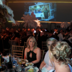 Attendees seated at a Foundation Ball dinner with place settings and glassware; two women in eveningwear pose at a table while palm trees and an illuminated stage screen showing photos of the Foundation's impact in the background.