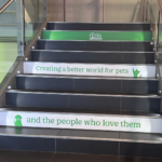 A stairwell with dark steps and glass railings, featuring green signage that reads “Pets” and “Creating a better world for pets and the people who love them.”