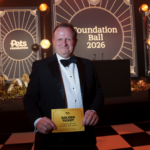 Man in a black tuxedo and bow tie stands on a checkered floor holding a Golden Ticket at the Pets Foundation Ball 2026 event, with a stage backdrop and warm string lights behind him.