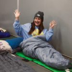 Woman in a blue sweatshirt and grey beanie smiling and raising hands while sitting in a sleeping bag indoors.