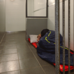 Person sleeping on a thin mattress with a pillow inside a small, bare room with tiled floor and metal bars nearby.