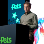 William Hewish stands behind a dark podium at a Pets Supplier Conference, with a “Pets” logo and star graphics on the backdrop and a large screen showing colourful pet imagery.