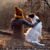 A woman with a brown bobble hat and glove with her arm around a white setter dog with brown ears, sitting on the ground. She has her back turned to the camer and the dog's head is turned looking a her.