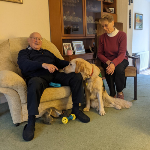 A man sitting in a chair with a yellow labrador dog sitting at his feet with his head in his lap. And a woman sitting in a chair next to the dog.